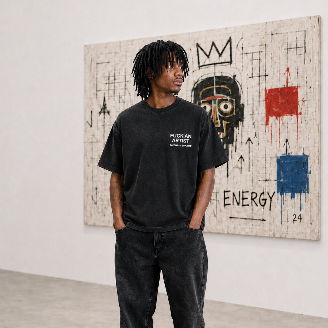 A man stands in a gallery wearing a black graphic t-shirt with white text that reads 'FUCK AN ARTIST' and 'ENERGY' on the pants, paired with black high-top sneakers featuring red accents. He is posed in front of a large abstract art print on a white wall.