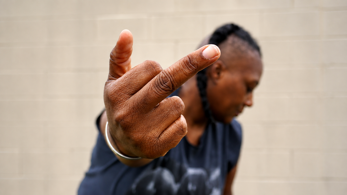 A close-up shot of a person's hand making a middle finger gesture, with their face blurred in the background against a light-colored brick wall.